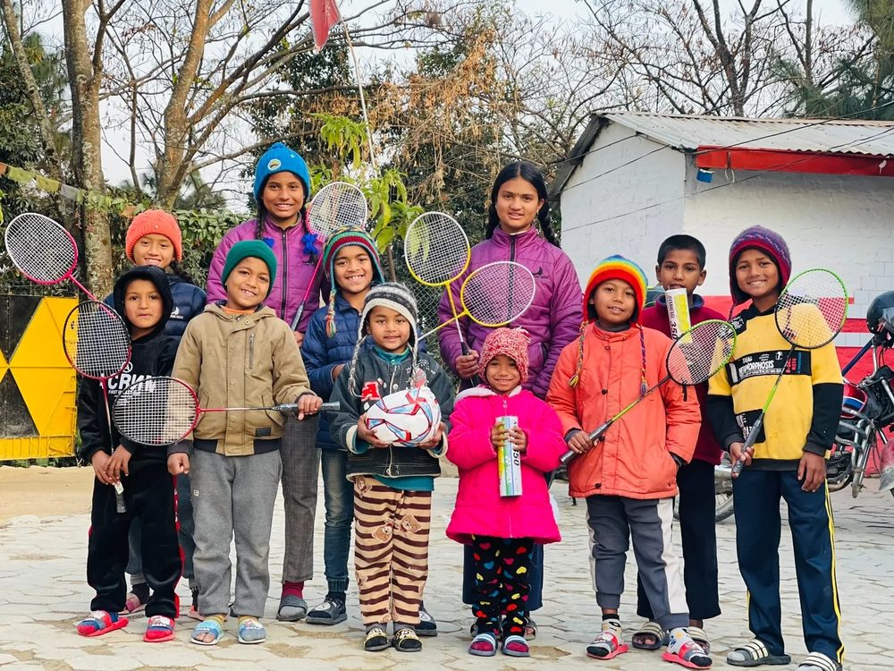 A group of children at the home outdoors in winter coats, holding badminton rackets and a ball.